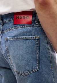 Κοντινό πλάνο στην τσέπη τζιν ανοιχτού μπλε χρώματος με ορατή πορτοκαλί ραφή και κόκκινη ετικέτα της μάρκας HUGO στη μέση κάτω από λευκό πουκάμισο.