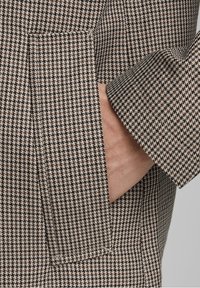 Ruka částečně v kapse rukávu blazeru s houndstooth vzorem v hnědé a krémové barvě, a viditelná postranní kapsa.