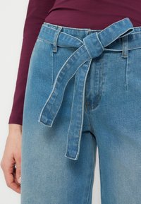 Γυναίκα που φοράει ανοιχτό μπλε τζιν ψηλής μέσης με ζώνη που δένει μόνη της και μπορντό μπλούζα με μακριά μανίκια, με το χέρι ακουμπισμένο δίπλα.