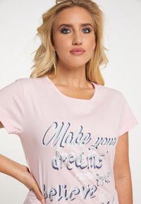 Helles rosa T-Shirt mit kurzen Ärmeln, mit strukturiertem weißen und blauen Drucktext: „Verwirkliche deine Träume.“ Weicher Stoff.