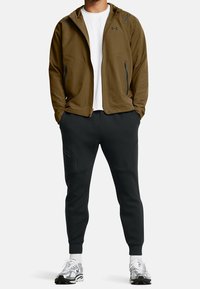 Mans atletiska outfit med en brun jacka med dragkedjefickor och en vit T-shirt under, tillsammans med svarta joggers och sneakers.