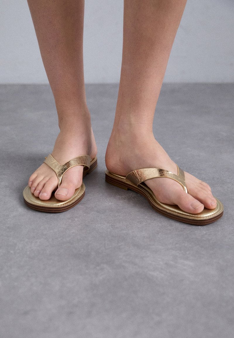 Gullflip-flops i gull med en teksturert stropp og lagdelt såle, med en liten bue og åpen tå-design. Flat brun base for stabilitet.