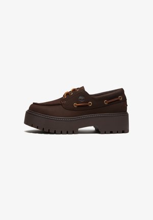 Chaussure bateau en cuir brun foncé avec semelle à crampons épaisse, détails en lacets, œillets métalliques et petit logo arbre sur le côté.