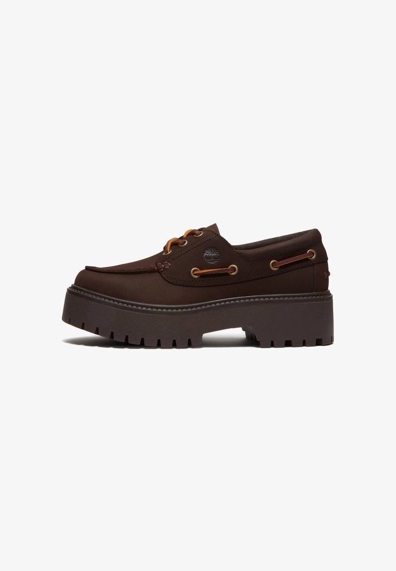 Chaussure bateau en cuir brun foncé avec semelle à crampons épaisse, détails en lacets, œillets métalliques et petit logo arbre sur le côté.