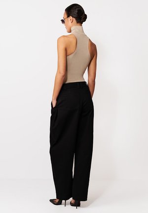 Kobieta z włosami spiętymi w kok, ubrana w bezrękawnik beżowy golf, szerokie czarne spodnie i okulary przeciwsłoneczne, stojąca z rękami w kieszeniach, ubrana w buty na wysokim obcasie.