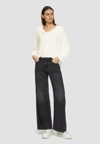 Chemisier blanc texturé avec un col en V et des détails en boutons, associé à un jean large sombre et des baskets blanches. Le mannequin se tient les mains dans les poches.