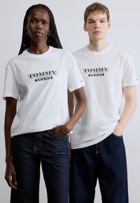 Λευκά βαμβακερά T-shirts με μεγάλο λογότυπο "TOMMY JEANS" σε μαύρο και γκρι. Συνοδεύονται από σκούρα μπλε τζιν παντελόνια και στα δύο μοντέλα.