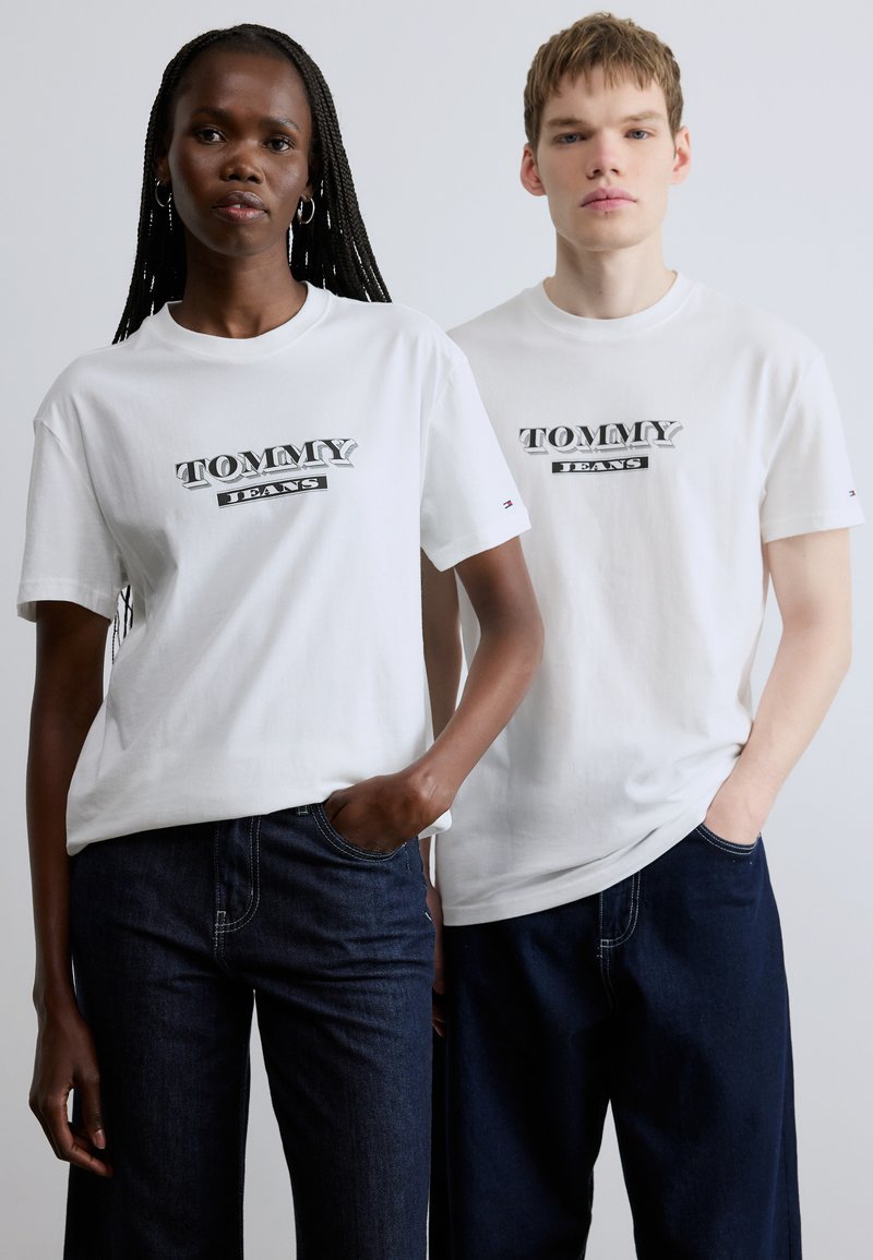 Λευκά βαμβακερά T-shirts με μεγάλο λογότυπο "TOMMY JEANS" σε μαύρο και γκρι. Συνοδεύονται από σκούρα μπλε τζιν παντελόνια και στα δύο μοντέλα.