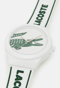 White silicone watch with green crocodile logo, white face, silver hour and minute hands, and green "LACOSTE" text on strap.