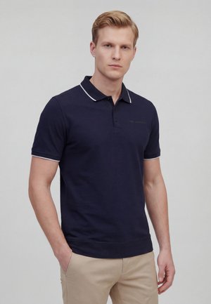 Blond man wearing a navy blue polo shirt with white trim and beige pants, standing with one hand in pocket against a plain background.