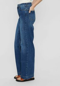 Bredbeniga blå denimjeans med hög midja, fyra fickor och rå kant, matchade med bruna platta sandaler.