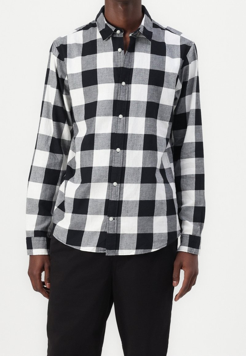 Homme portant une chemise à manches longues à carreaux noirs et blancs avec un pantalon noir, debout devant un fond blanc uni.