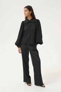 Black, textured blouse with a button front, ruffled collar, and voluminous sleeves, paired with tailored black wide-leg trousers and sandals.