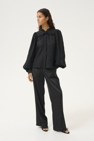 Zwarte, textuur blouse met knoopsluiting, gerimpelde kraag en volumineuze mouwen, gecombineerd met op maat gemaakte zwarte wijde broek en sandalen.