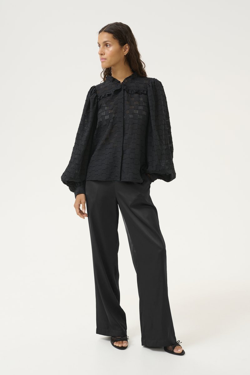 Black, textured blouse with a button front, ruffled collar, and voluminous sleeves, paired with tailored black wide-leg trousers and sandals.