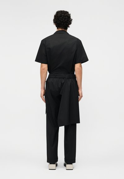 Muž s kudrnatými vlasy otočený zády, oblečený v černém krátkém rukávu, černých kalhotách s bočními přehyby látky a černo-bílých botách.