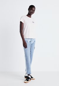 Femme portant un t-shirt rose pâle, un jean bleu clair avec des détails de couture à l'avant, des chaussettes blanches et des baskets noir et blanc, se tenant devant un fond blanc uni.