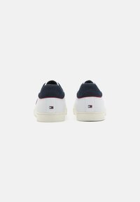 Zapatillas blancas con un cuello de punto azul marino, acentos rojos, suelas de goma planas y un pequeño parche con el logo en el talón. Textura de cuero suave.