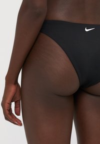 Svart bikini-bunn med høy skjæring, med en liten hvit Nike-logo på baksiden. Slank, glatt stoff med minimale sømmer.