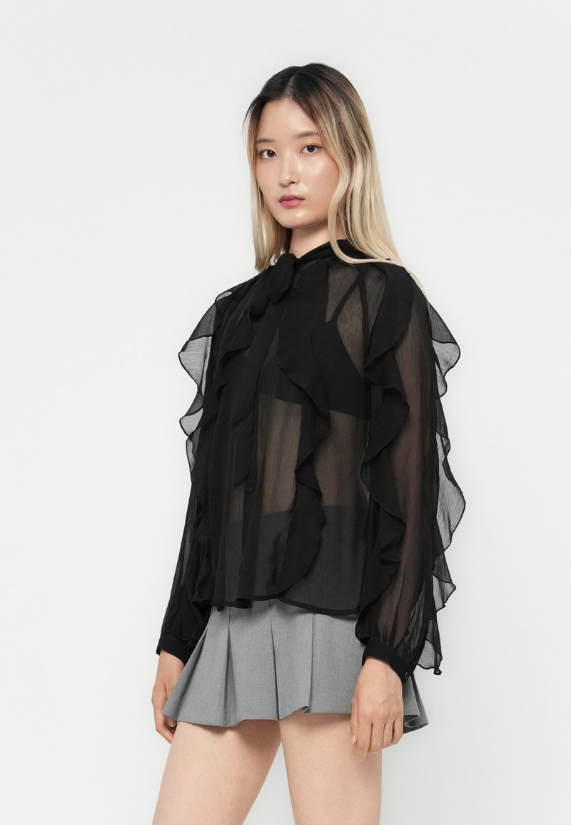 Blouse noire transparente avec des détails froncés, fermeture à boutons et un nœud au cou ; associée à une jupe plissée gris clair.