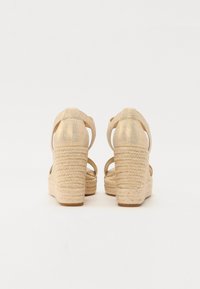 Sandales à talon compensé beige avec une plateforme en jute texturé et des brides métalliques au niveau de la cheville. Design à dos ouvert et détails tissés à motifs sur le talon.