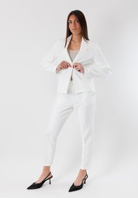 Completo bianco composto da blazer e pantaloni sartoriali, con una texture liscia. Il blazer presenta un colletto a punta, mentre i pantaloni hanno una vestibilità slim. Indossato con tacchi neri.