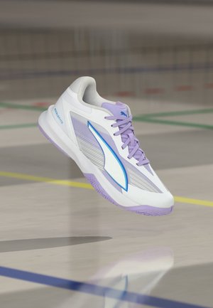 Άσπρο και λεβαντί αθλητικό παπούτσι Puma με μπλε λογότυπο να αιωρείται πάνω από πάτωμα εσωτερικού αθλητικού γηπέδου.
