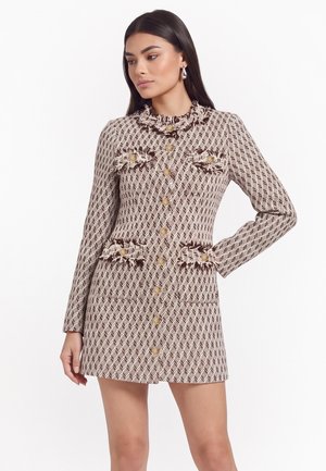 Donna con lunghi capelli scuri che indossa un vestito cappotto a fantasia con bottoni dorati e tasche con frange, posando con una mano sui fianchi.