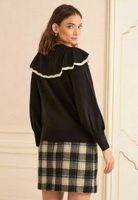 Pulover negru cu un guler volănat în crem, dintr-un material texturat, și mâneci bufante, asortat cu o fustă în carouri în tonuri de bej și negru.