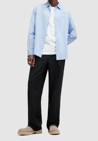 Uomo che indossa una camicia azzurro chiaro sopra una t-shirt bianca, pantaloni neri, stivali in suede beige e un anello d'argento sulla mano sinistra.
