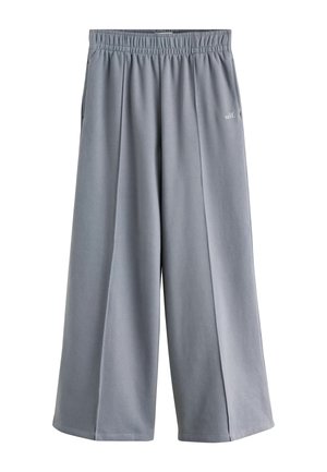 Blonde Frau trägt weißes kurzärmeliges Hemd und graue weite Hose, steht draußen mit Händen in den Taschen und hält eine braune Tasche.
