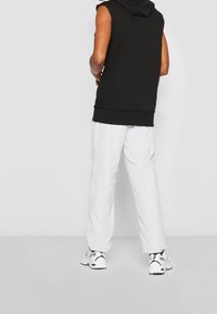 Black sleeveless hoodie with a textured finish, paired with loose white pants, and white sneakers with black accents.