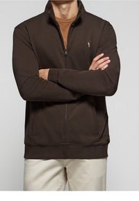 Brun zip-jacka i ett mjukt tyg, med hög krage, sidofickor och en subtil logotyp på vänster bröst.