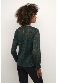 Blusa verde in pizzo con un motivo geometrico, maniche lunghe con polsini arricciati e schiena con apertura a chiave dotata di chiusura a bottone.