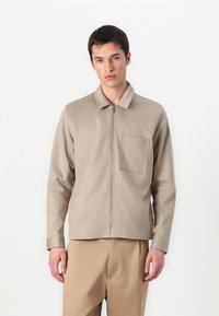 Bežinė zip-up striukė su apykakl�ėmis, ilgomis rankovėmis ir krūtinės kišene. Minkšta medžiaga su lygiu paviršiumi. Kelnės atitinkamos spalvos.