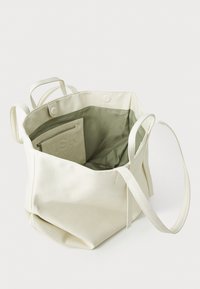 Sac fourre-tout blanc, texturé, avec un haut ouvert et deux poignées. Dispose d'un intérieur doublé vert clair avec une poche zippée.