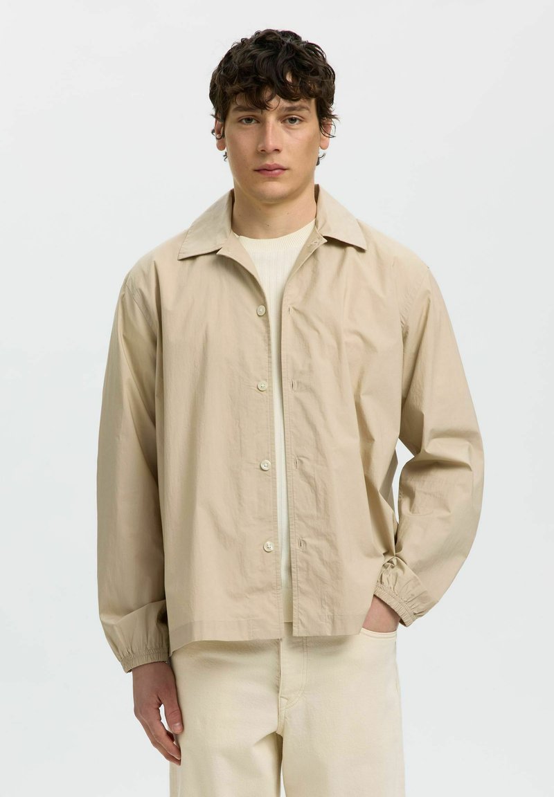 Jeune homme aux cheveux bouclés foncés portant une chemise beige boutonnée par-dessus un pull clair et un pantalon crème, debout devant un fond blanc uni.