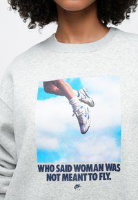 Γκρι φούτερ με σχέδιο μπρ οστά που απεικονίζει ένα πόδι με λευκά και μπλε αθλητικά παπούτσια πάνω σε μπλε ουρανό, με το κείμενο: "ΠΟΙΟΣ ΕΙΠΕ ΟΤΙ Η ΓΥΝΑΙΚΑ ΔΕΝ ΠΡΕΠΕΙ ΝΑ ΠΕΤΑ".
