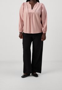 Blusa rosa chiaro con scollatura a V, maniche a sbuffo e cuciture decorative. Abbinata a pantaloni neri a gamba larga e ballerine nere.