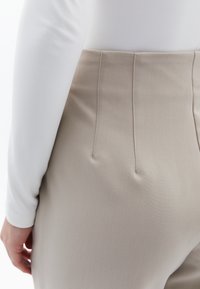 Beige hoge taille broek van een gestructureerde stof, met drie verticale plooien aan de achterkant en een smooth witte blouse met lange mouwen.