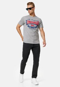 Grå bomulds t-shirt med rød og blå grafisk tekst "The Original Lonsdale Since 1960." Parret med mørke jeans og hvide sneakers.