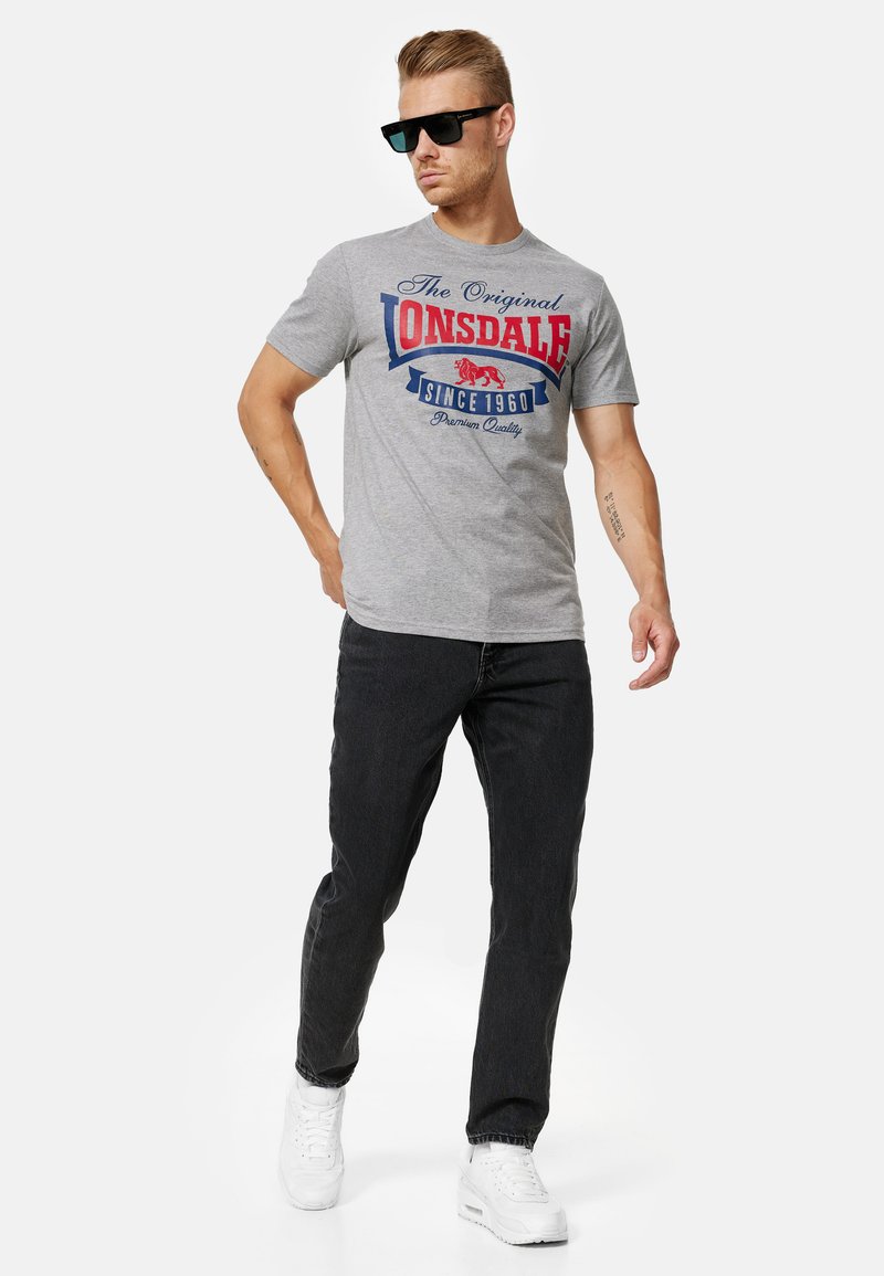 Grå bomulds t-shirt med rød og blå grafisk tekst "The Original Lonsdale Since 1960." Parret med mørke jeans og hvide sneakers.