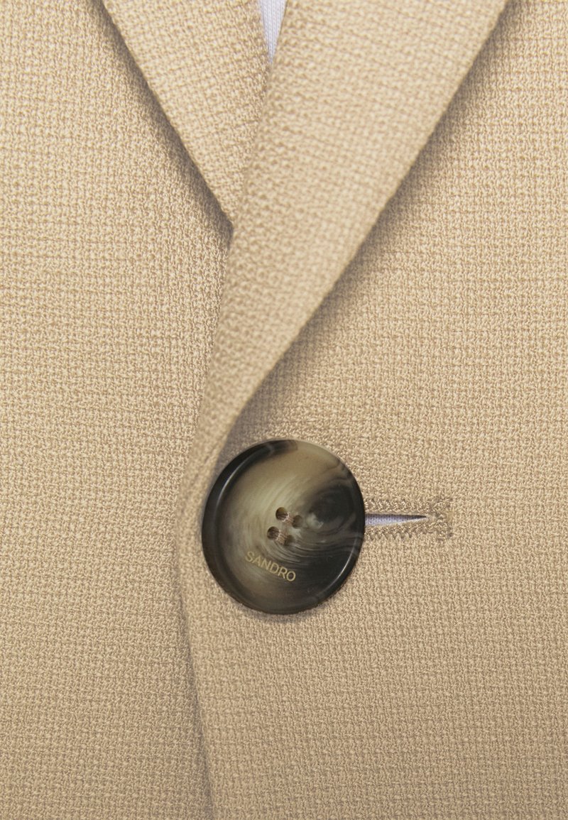 Beige textured blazer featuring a round, dark button with a swirling pattern and "SANDRO" engraved. Button secures left lapel.
