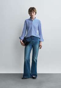 Persoană în picioare purtând cămașă albastră și albă cu dungi verticale, blugi evazați albaștri, pantofi negri, ținând o geantă plic neagră pe un fundal simplu.