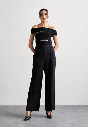 Zwarte off-shoulder top met uitsnijdingen, gecombineerd met zwarte high-waisted wijde broek en zwarte puntige hakken. Gladde stoftextuur.