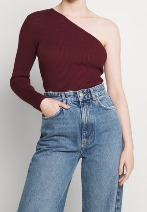 Osoba ubrana w prążkowany, bordowy sweter na jedno ramię z długim rękawem i jasnoniebieskie, wysokowyższe dżinsy.