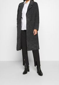 Cappotto lungo, grigio scuro, con texture, a due bottoni e tasche laterali, indossato sopra una camicia bianca e pantaloni neri, completato da stivaletti neri con tacco.