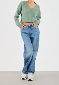 Pull court vert clair en maille texturée associé à un jean bleu taille haute. Le mannequin porte des baskets claires, debout devant un fond neutre.