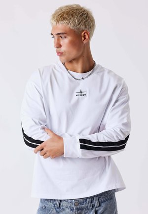 Joven con el pelo rubio corto, lleva una camisa blanca de manga larga con rayas negras y un collar de cadena, mirando de lado.