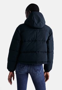 Mörkblå puffjacka med huva, med horisontella quiltade sektioner och elastiska ärmslut. Bärs över mörka jeans.